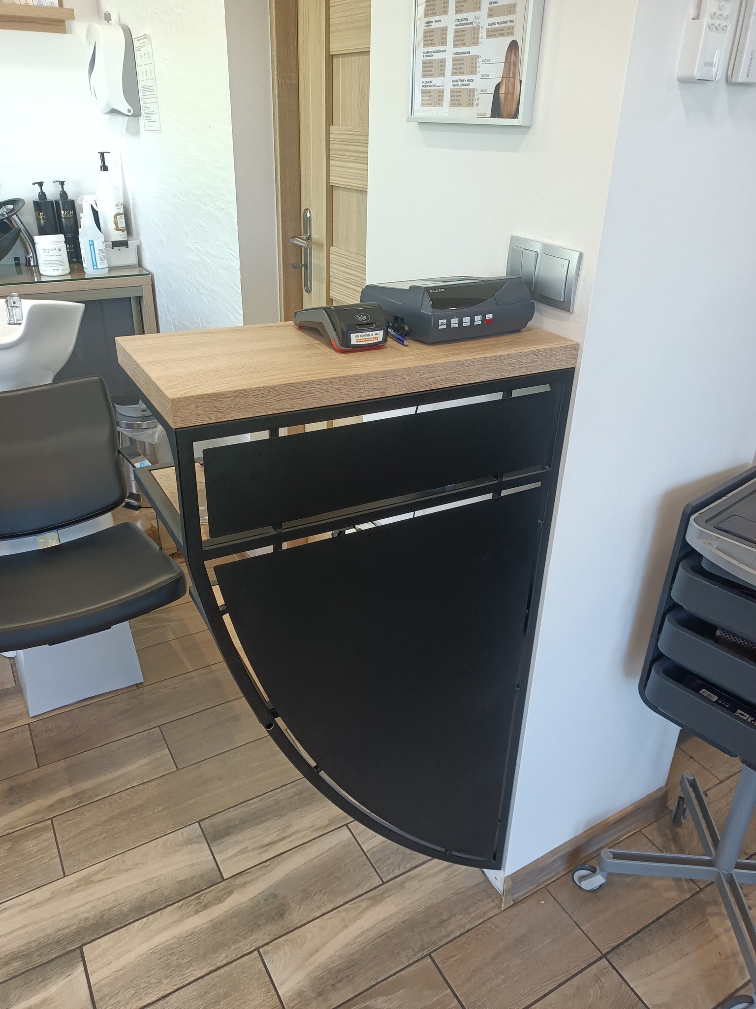 Stylowa lada recepcyjna z drewnianym blatem i czarną, metalową konstrukcją o zaokrąglonym kształcie, wyposażona w terminal płatniczy i kasę fiskalną w salonie fryzjerskim.