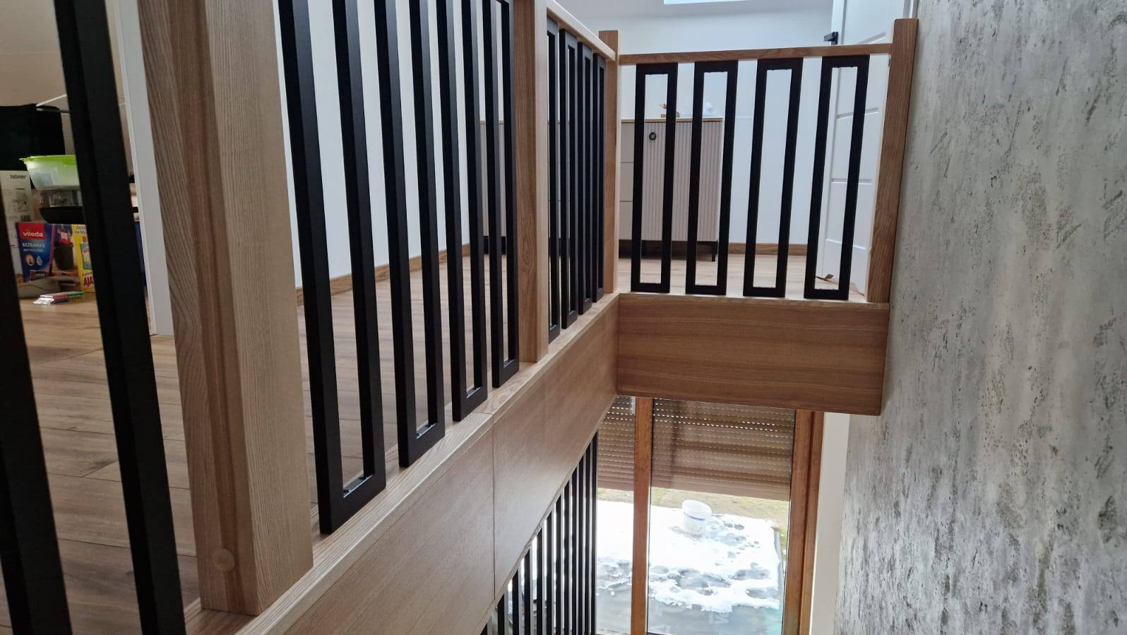 Nowoczesna balustrada schodowa z czarnymi, pionowymi elementami i drewnianymi słupkami, widok z góry, jasne wnętrze z oknem w tle.