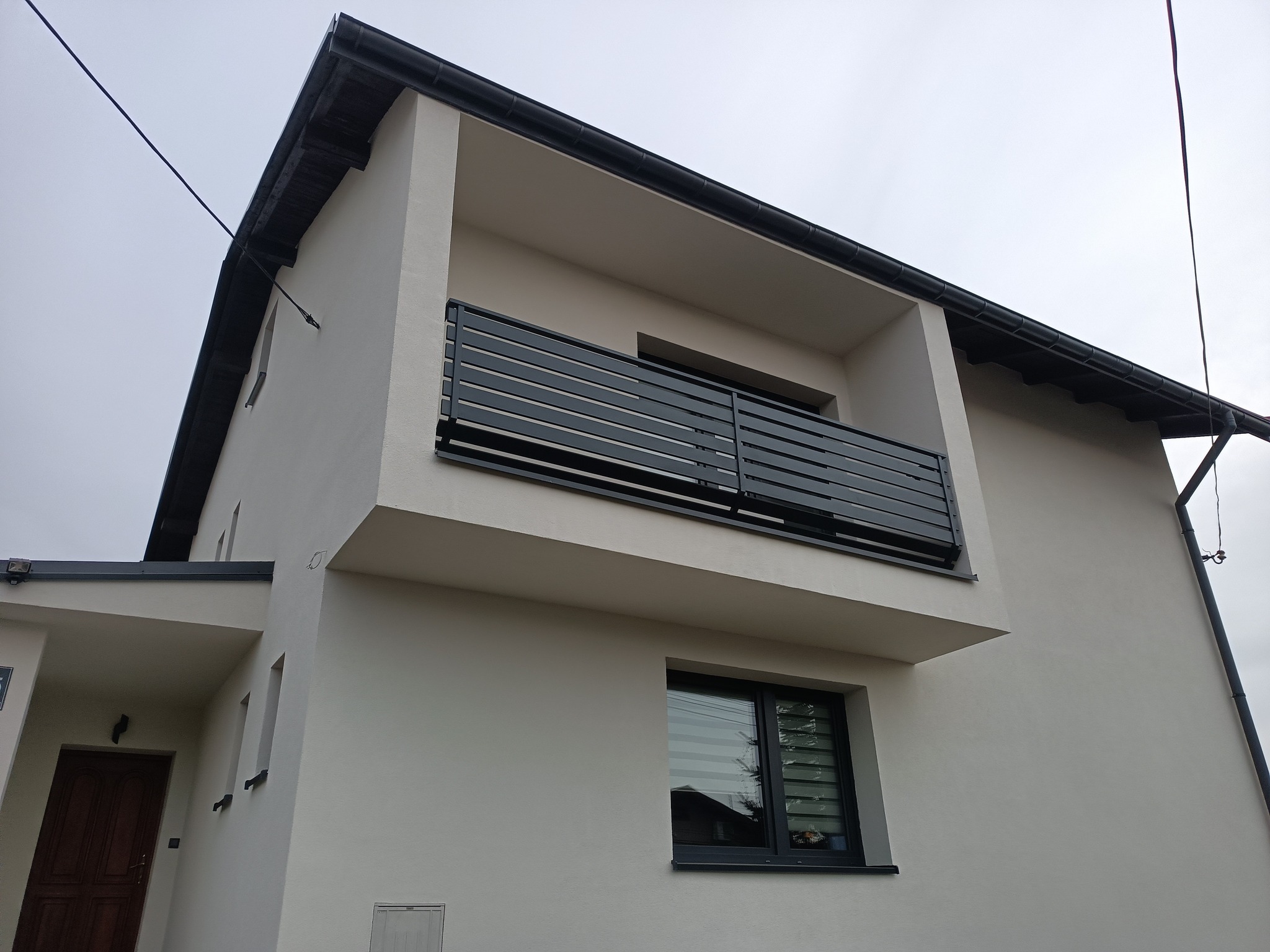 Nowoczesny balkon z czarną, poziomą balustradą na tle jasnej elewacji domu. Minimalistyczny design, proste linie, widok z dołu, pochmurne niebo.