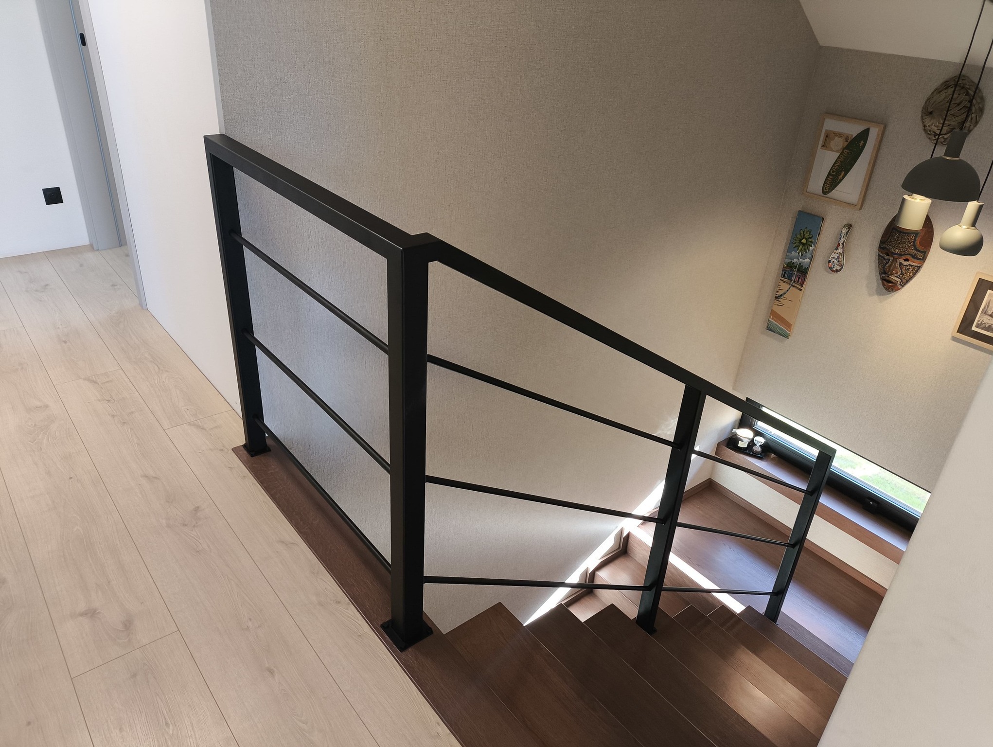 Nowoczesne schody z drewnianymi stopniami i czarną, minimalistyczną balustradą z wypełnieniem z siatki. Widok z góry, jasne wnętrze z dekoracjami na ścianie.