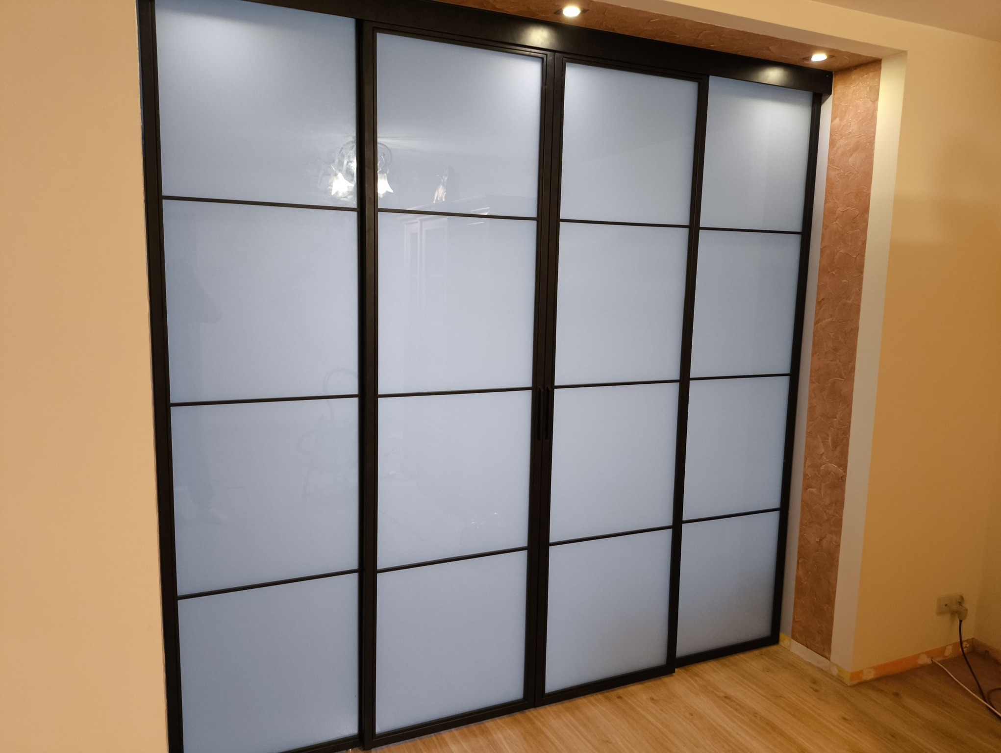 Nowoczesna zabudowa szafy przesuwnej z czarnymi profilami i mlecznym szkłem, widoczna w jasnym wnętrzu. Minimalistyczny design, podkreślony przez subtelne oświetlenie.