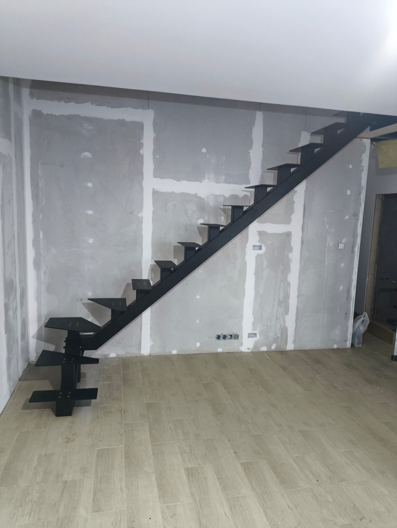 Nowoczesne, czarne schody metalowe bez balustrady, zamontowane w surowym wnętrzu z widocznymi śladami prac wykończeniowych na ścianach. Minimalistyczny design.