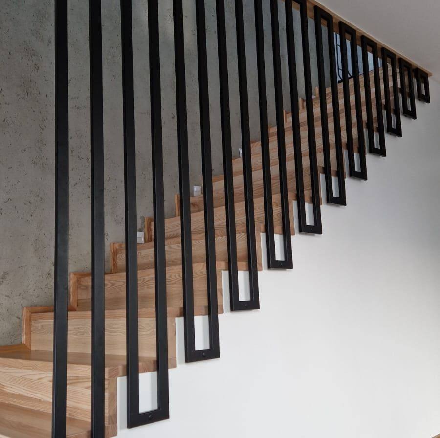 Drewniane schody z czarną, minimalistyczną balustradą z pionowych, prostokątnych elementów. Betonowa ściana w tle. Nowoczesny design.