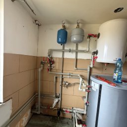 Damir Khassan - Złożona instalacja grzewcza z dwoma zbiornikami wyrównawczymi, bojlerem i licznymi rurami z zaworami. Widoczne narzędzia i butelka wody. Ściany wyłożone częściowo kafelkami.