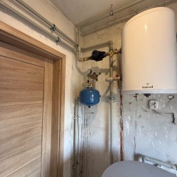 Damir Khassan - Bojler Galmet z osprzętem i rurami w surowym pomieszczeniu. Widoczne ślady na ścianie i fragment drzwi. Instalacja grzewcza w trakcie montażu.