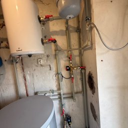 Damir Khassan - Biały bojler Galmet z szarym zbiornikiem wyrównawczym, połączony rurami z zaworami Ferro, zamontowany na ścianie z widocznymi śladami po instalacji.