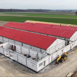 INHOL CONSTRUCTION SPÓŁKA Z OGRANICZONĄ ODPOWIEDZIALNOŚCIĄ - Widok z lotu ptaka na trzy równoległe budynki gospodarcze w trakcie budowy: dwa z czerwonym pokryciem dachowym, jeden z widoczną drewnianą więźbą. Widać maszyny budowlane i pracowników na placu.