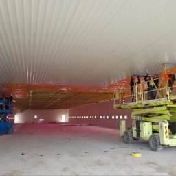 INHOL CONSTRUCTION SPÓŁKA Z OGRANICZONĄ ODPOWIEDZIALNOŚCIĄ - Montaż białych paneli sufitowych i pomarańczowej izolacji termicznej w przestronnej hali przemysłowej. Pracownicy na dwóch podnośnikach nożycowych, widoczne kwadratowe otwory w ścianach.