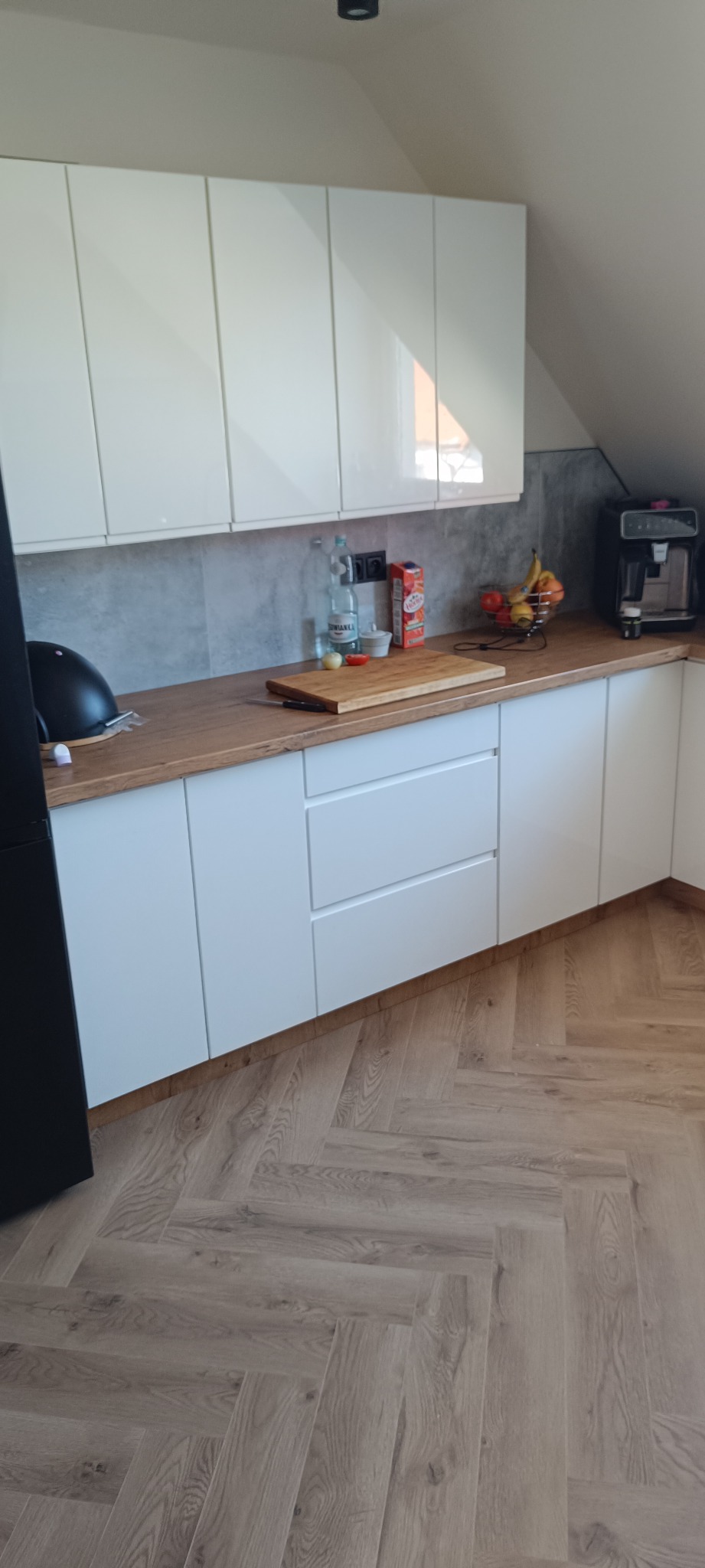 Jasna kuchnia na poddaszu z białymi szafkami, drewnianym blatem i podłogą w jodełkę. Na blacie deska do krojenia, owoce i ekspres do kawy.