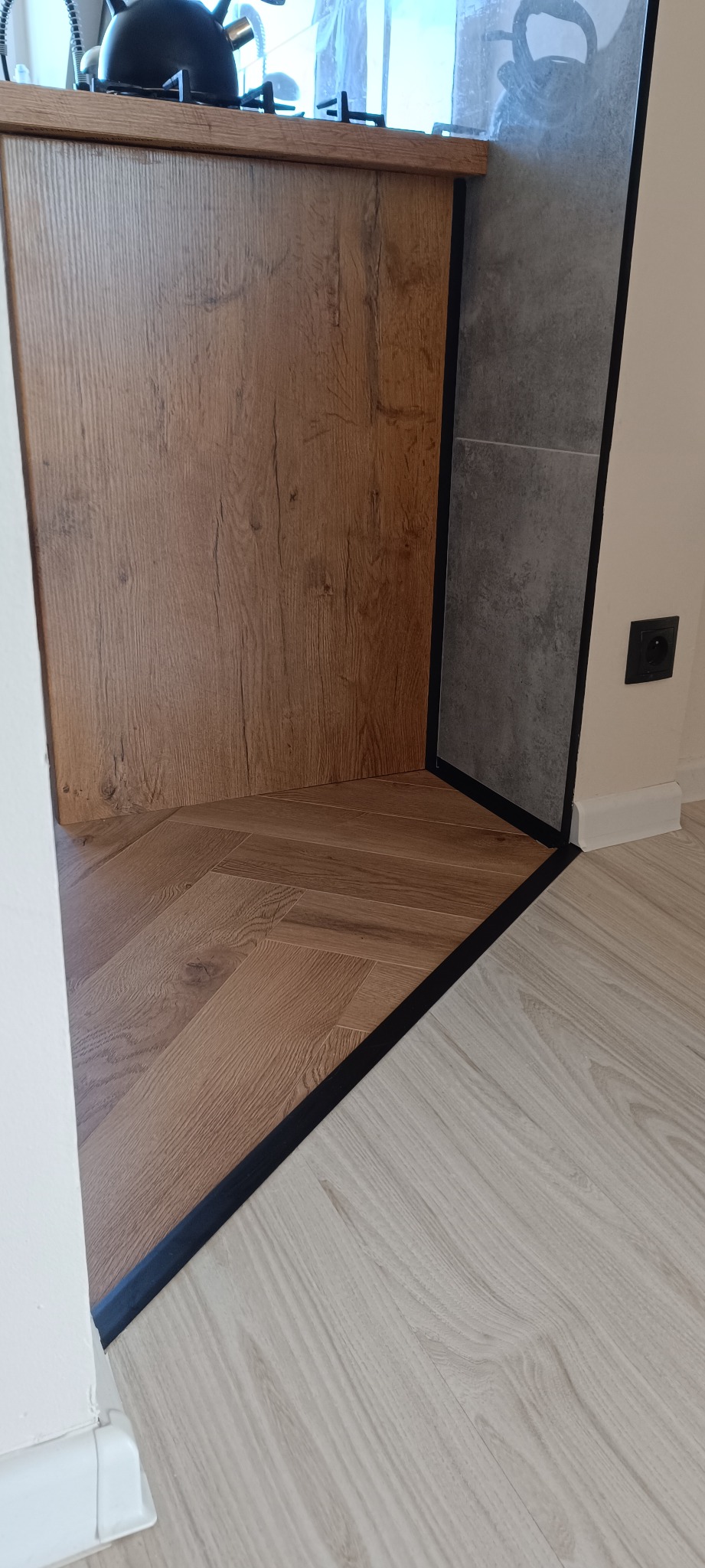 Połączenie dwóch rodzajów podłóg: parkiet ułożony w jodełkę i panele, oddzielone czarną listwą. Widoczny fragment zabudowy kuchennej i szare płytki na ścianie.