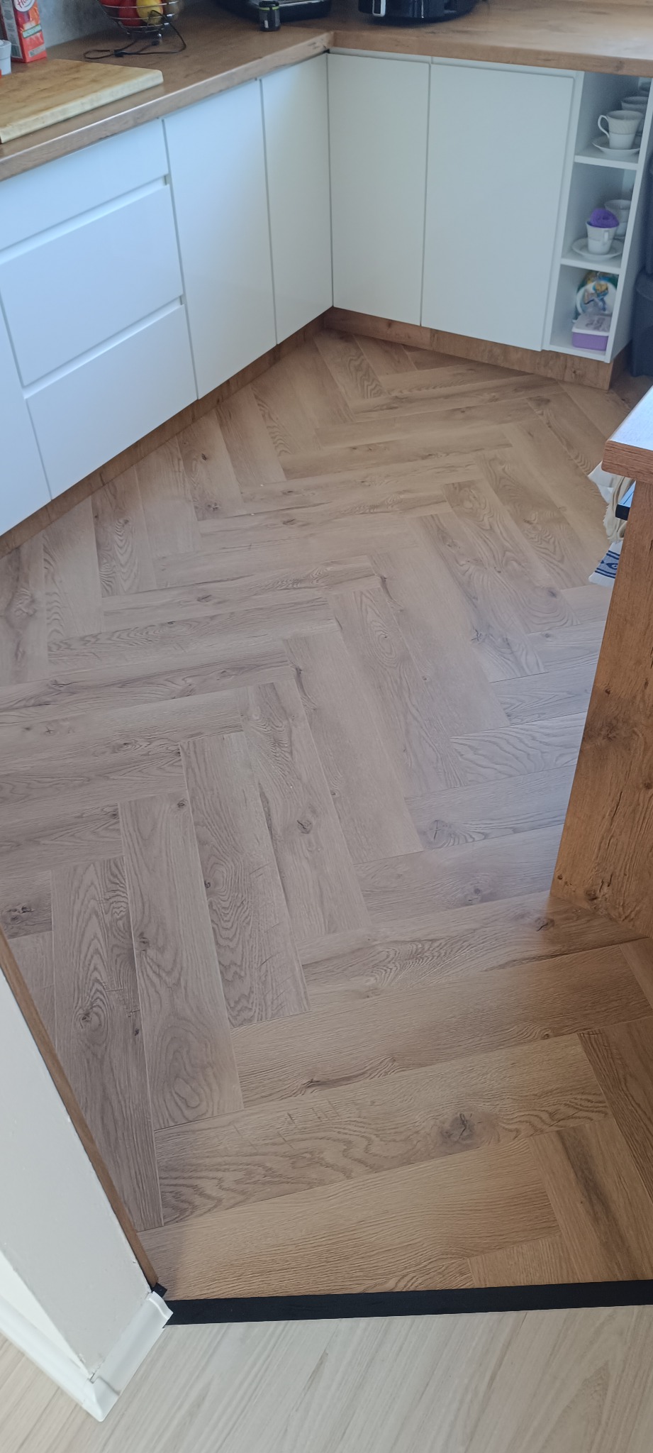 Ułożona podłoga z paneli w jodełkę w kuchni z białymi szafkami. Widoczny fragment progu i drewniany blat. Jasne, naturalne światło.