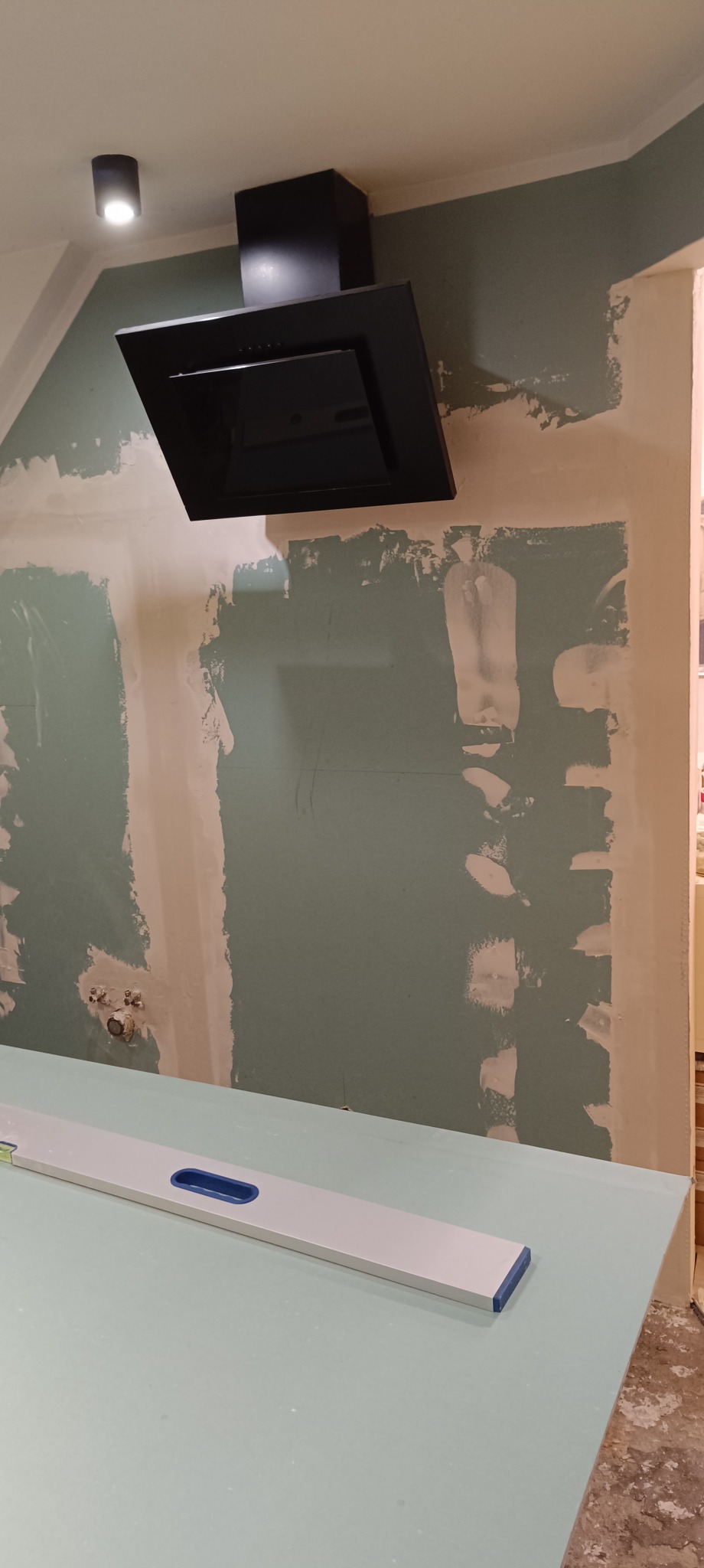 Kuchnia w trakcie prac: czarny okap nad płytą karton-gips, ściana z widocznymi śladami szpachlowania. Poziomica na blacie. W tle widoczny fragment podłogi.