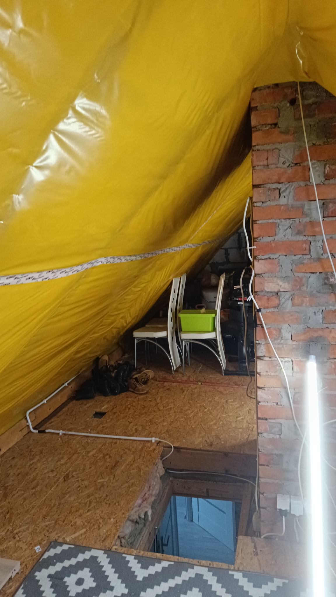 Poddasze z żółtą folią izolacyjną, fragmentem muru z cegły i drewnianą podłogą. Widoczne krzesła, przewody elektryczne i wejście na niższy poziom.
