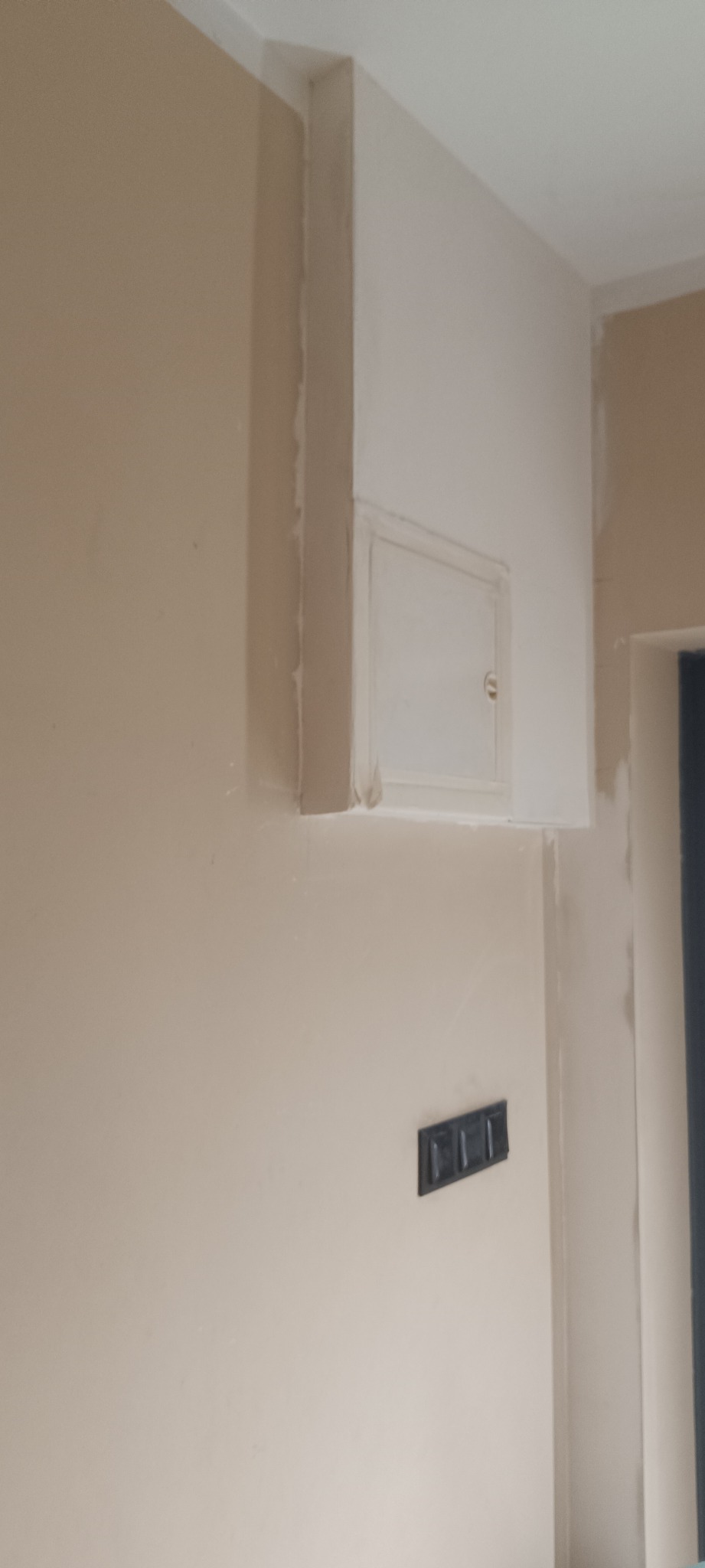 Ściana w trakcie wykańczania z widocznym otworem rewizyjnym i gniazdkiem elektrycznym. Widoczne ślady po szpachlowaniu i malowaniu.