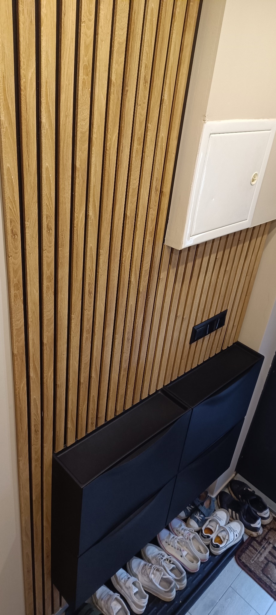 Nowoczesna aranżacja przedpokoju: drewniane lamele ścienne, czarna szafka na buty i biała klapa rewizyjna. Minimalistyczny design z dbałością o detale.