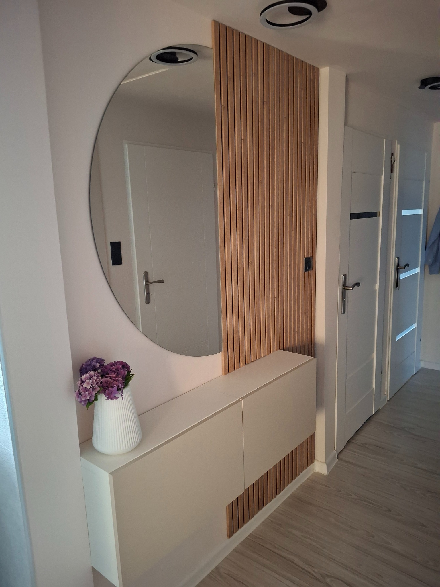 Nowoczesny przedpokój z okrągłym lustrem, lamelami dekoracyjnymi i białą szafką. Na szafce wazon z fioletowymi kwiatami. Widoczne białe drzwi.