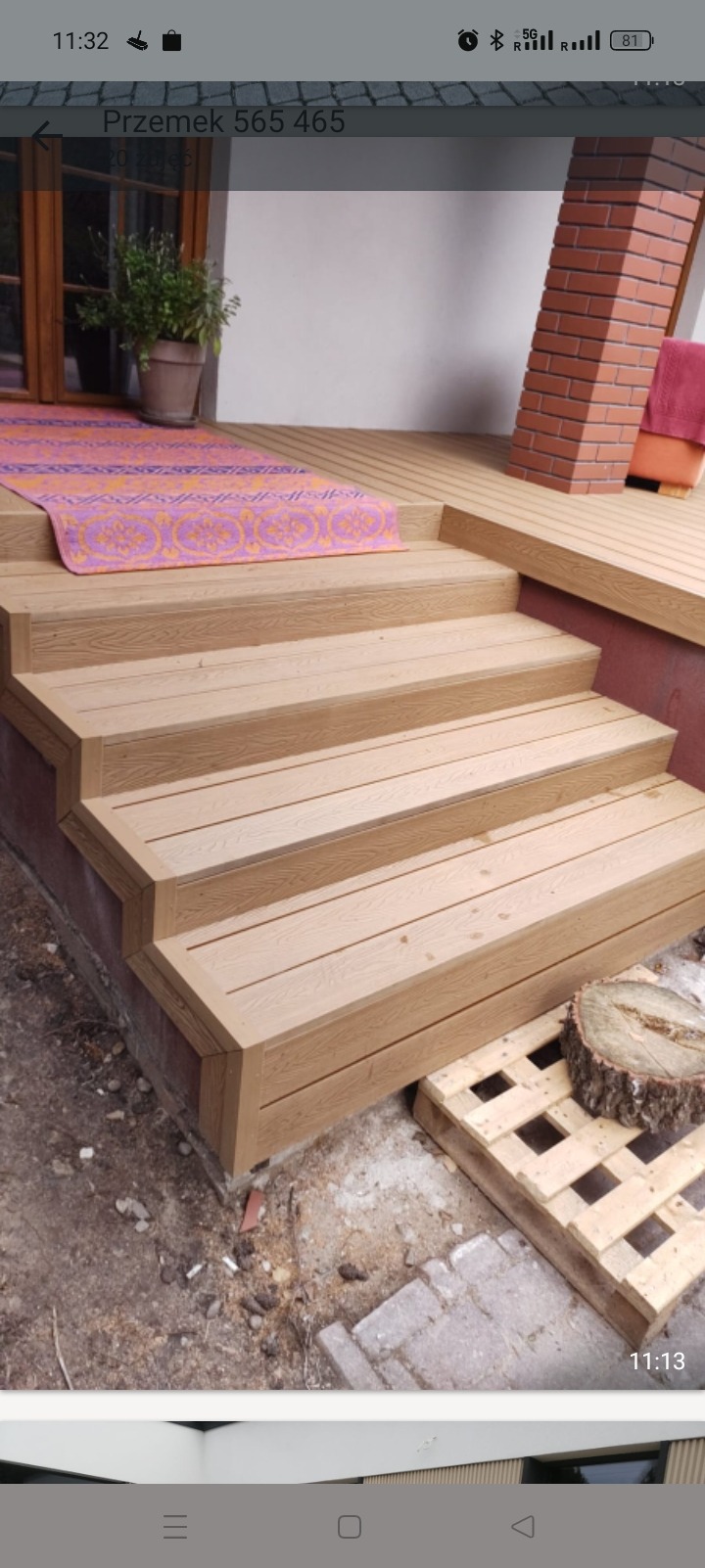 Schody z desek kompozytowych prowadzące na taras. Widoczny fragment tarasu, ceglany filar i dywan. Na dole paleta i pień drzewa. Ujęcie z dołu.