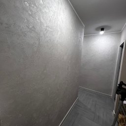 NaturaVerde - Korytarz po malowaniu ścian farbą dekoracyjną w odcieniach szarości z delikatnym połyskiem. Na podłodze szare płytki ułożone w jodełkę. Widoczne oświetlenie sufitowe.