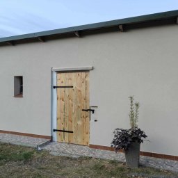 NaturaVerde - Otynkowany budynek gospodarczy z drewnianymi drzwiami i małym oknem. Obok drzwi stoi donica z rośliną. Widoczny fragment chodnika i trawnika.