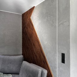 NaturaVerde - Stylowa ściana z drewnianą dekoracją i szarą fakturą, oświetlona listwą LED. Fragment szarej sofy z pledem w rogu kadru. Nowoczesny design wnętrza.