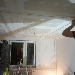 FIL-BUD Filip Labok - Sufit z płyt karton-gips, częściowo wyszpachlowany, z widocznymi przewodami elektrycznymi. W tle okno i drabina, narzędzia pracy. Wnętrze w trakcie prac wykończeniowych.