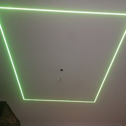 FIL-BUD Filip Labok - Podwieszany sufit z oświetleniem LED w kształcie trapezu, emitującym zielone światło. Widoczne przewody w centralnej części sufitu, sugerujące dalsze prace wykończeniowe.