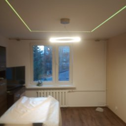 FIL-BUD Filip Labok - Pokój z drewnianą podłogą i oknem. Na suficie widoczna lampa w kształcie okręgu i zielony laser tworzący kwadrat. Meble zabezpieczone folią malarską.