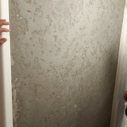 FIL-BUD Filip Labok - Dekoracyjna ściana z efektem betonu architektonicznego w odcieniach szarości, widoczna faktura i nieregularne wzory. Fragment ościeżnicy drzwi po lewej stronie.