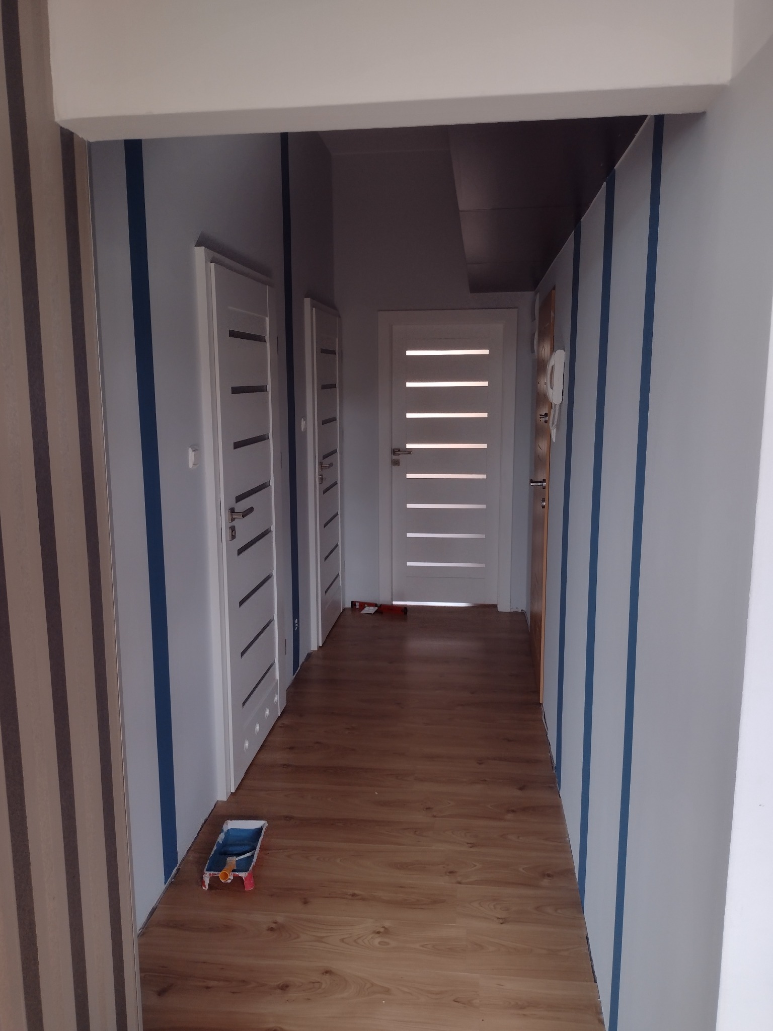 Korytarz po malowaniu ścian w biało-szare pasy z niebieskimi akcentami. Widoczne drzwi i podłoga z paneli. Na podłodze wałek malarski.