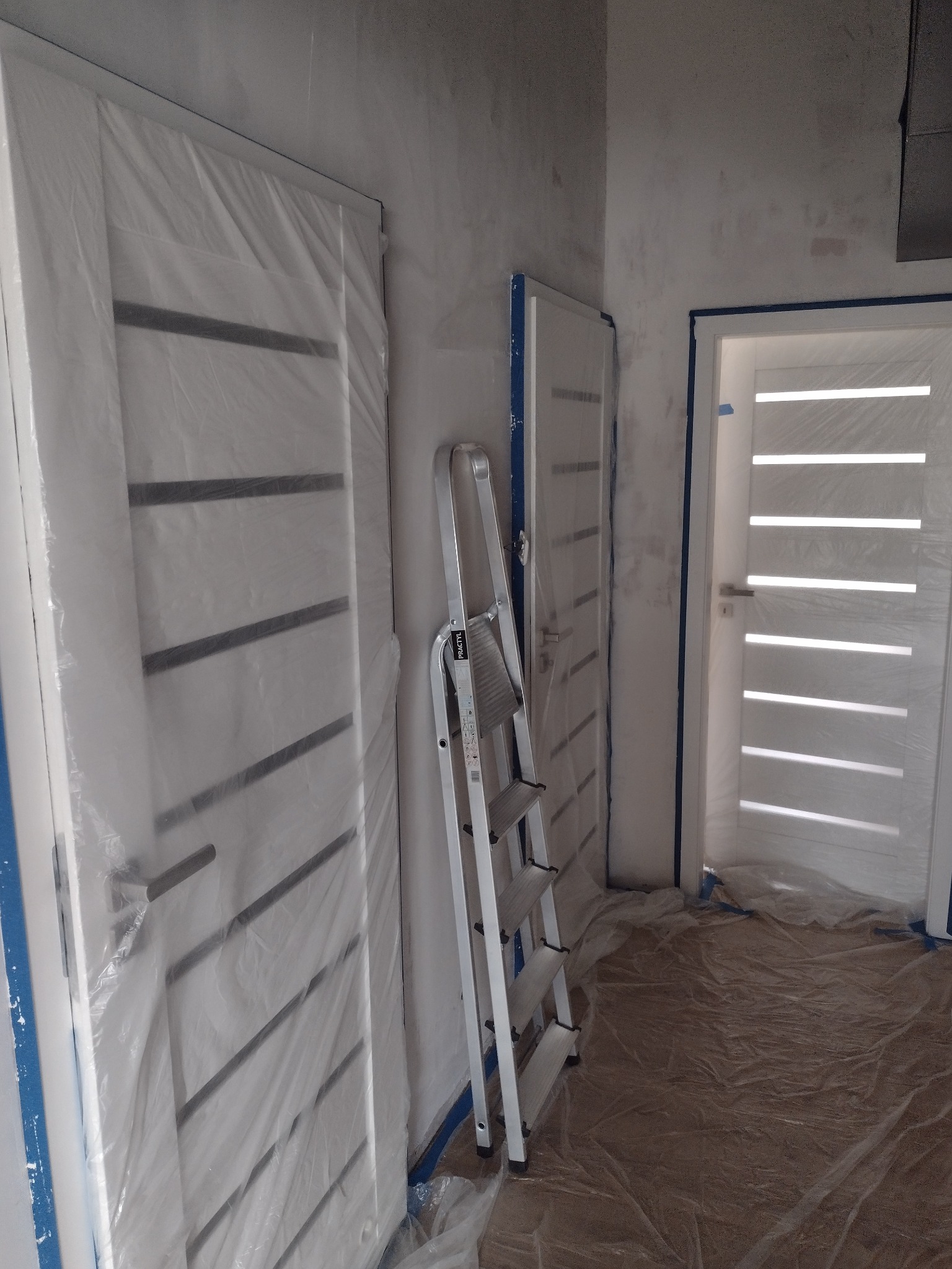 Białe, nowoczesne drzwi z poziomymi panelami, zabezpieczone folią malarską. Aluminiowa drabina oparta o ścianę. Przygotowania do malowania, podłoga zabezpieczona folią.