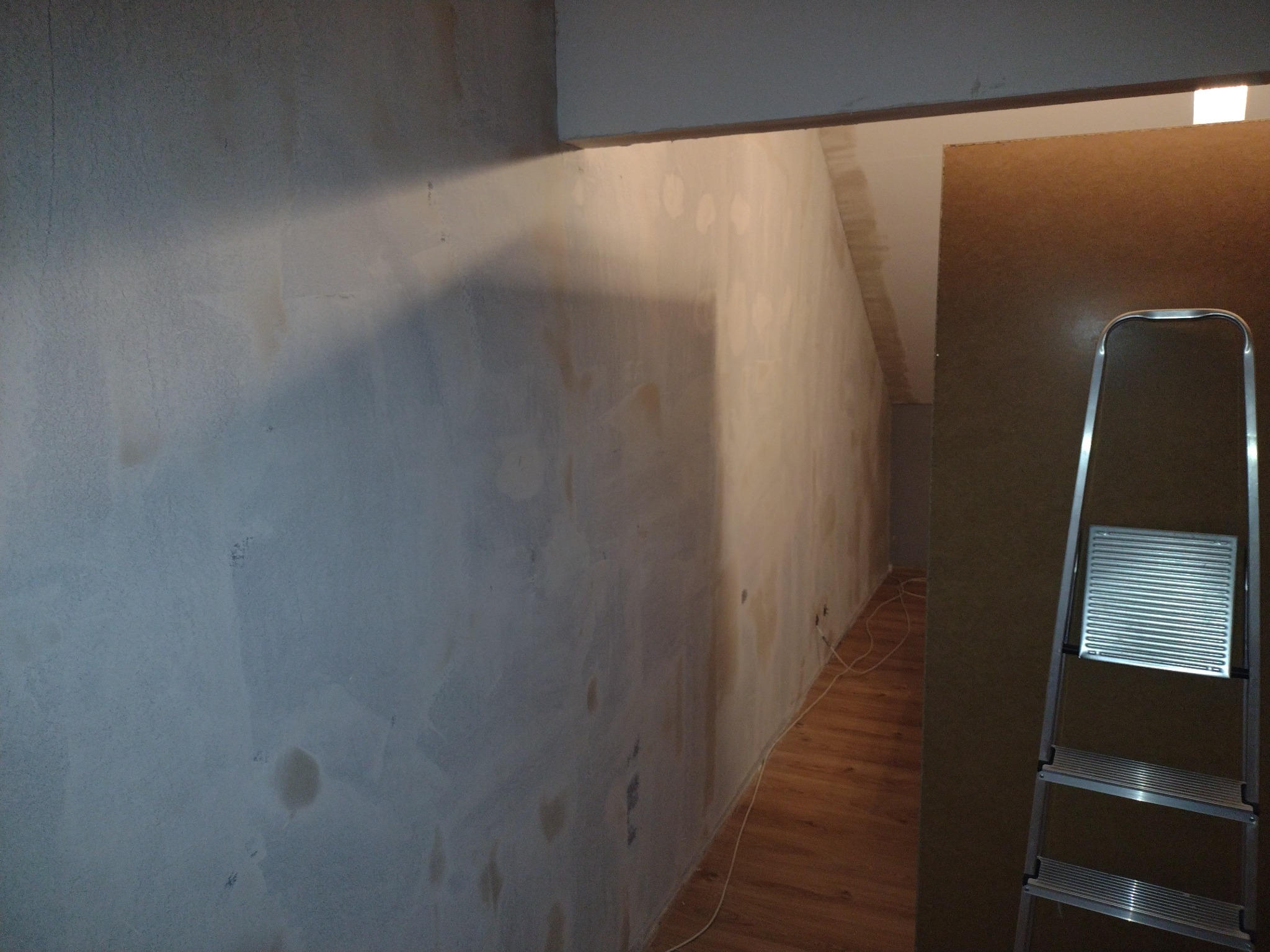 Ściana po szpachlowaniu, widoczne ślady po łatach, przygotowana do malowania. W rogu stoi drabina, a w tle widać podłogę z paneli.