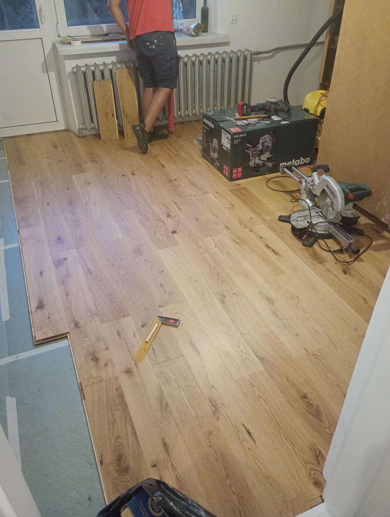 Układanie paneli podłogowych w jasnym odcieniu drewna. Widoczna piła do cięcia paneli, miarka kątowa i częściowo ułożona podłoga. W tle osoba i grzejnik.