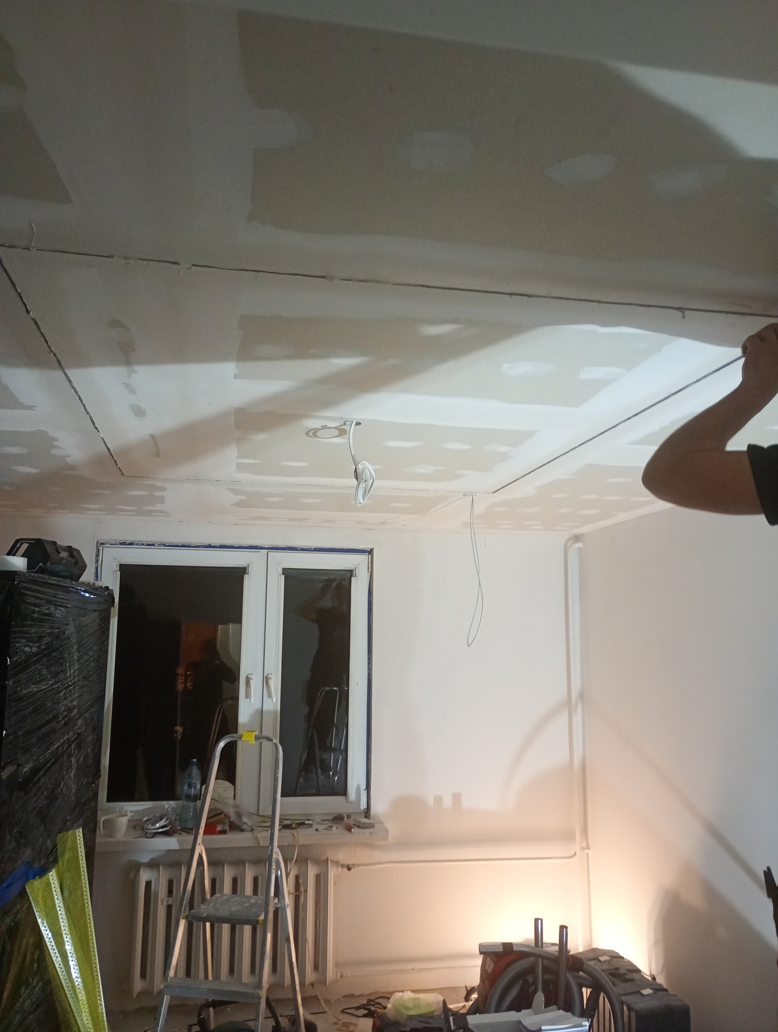 Sufit z płyt karton-gips, częściowo wyszpachlowany, z widocznymi przewodami elektrycznymi. W tle okno i drabina, narzędzia pracy. Wnętrze w trakcie prac wykończeniowych.