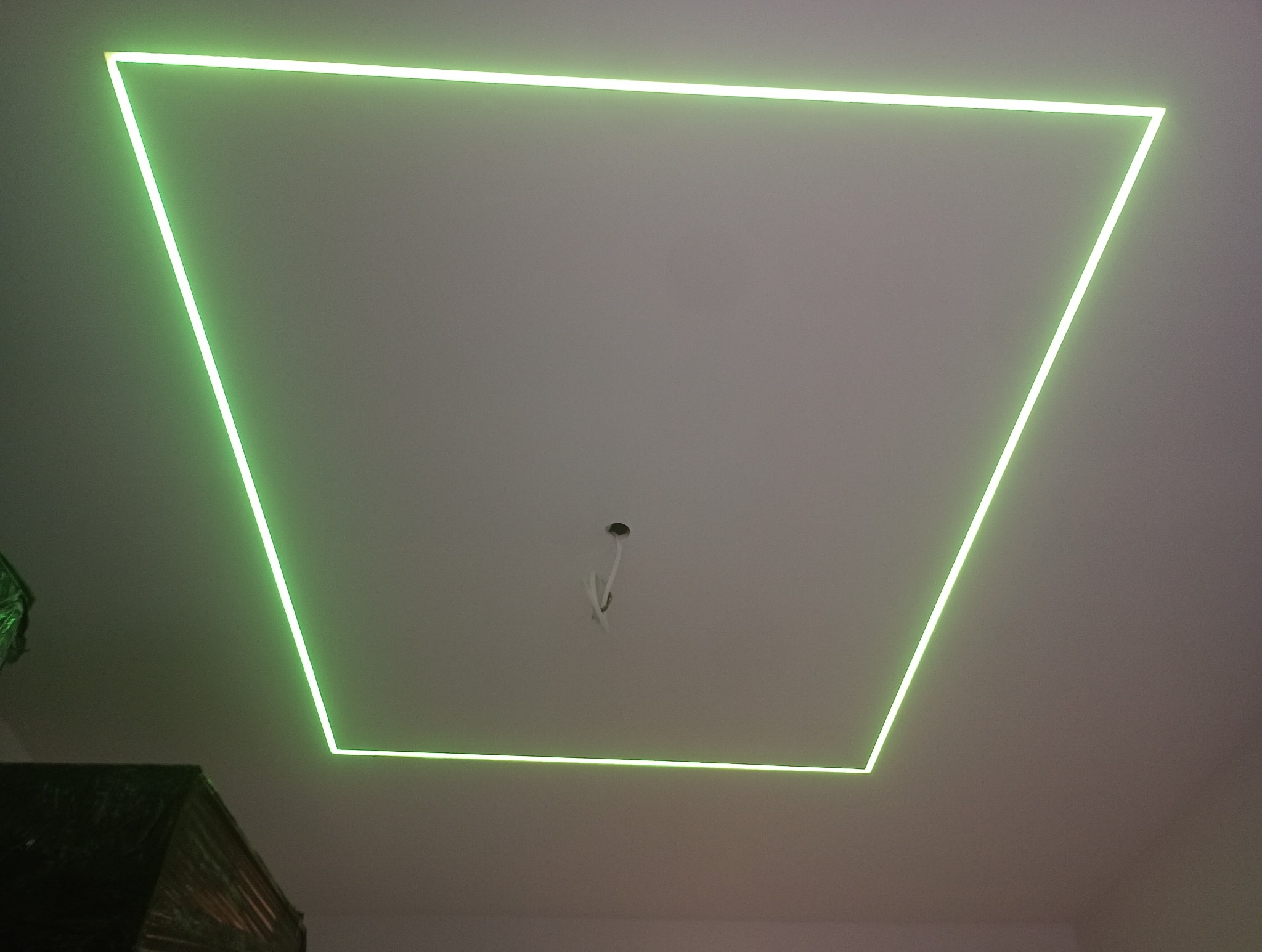 Podwieszany sufit z oświetleniem LED w kształcie trapezu, emitującym zielone światło. Widoczne przewody w centralnej części sufitu, sugerujące dalsze prace wykończeniowe.