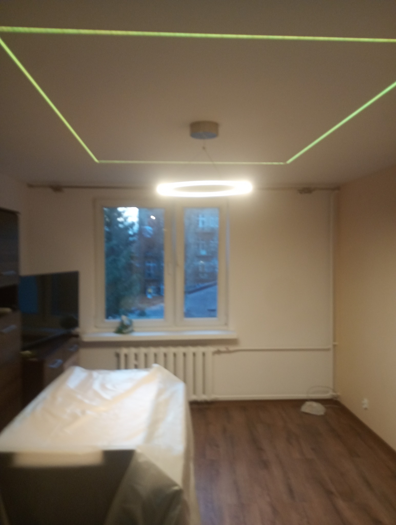 Pokój z drewnianą podłogą i oknem. Na suficie widoczna lampa w kształcie okręgu i zielony laser tworzący kwadrat. Meble zabezpieczone folią malarską.