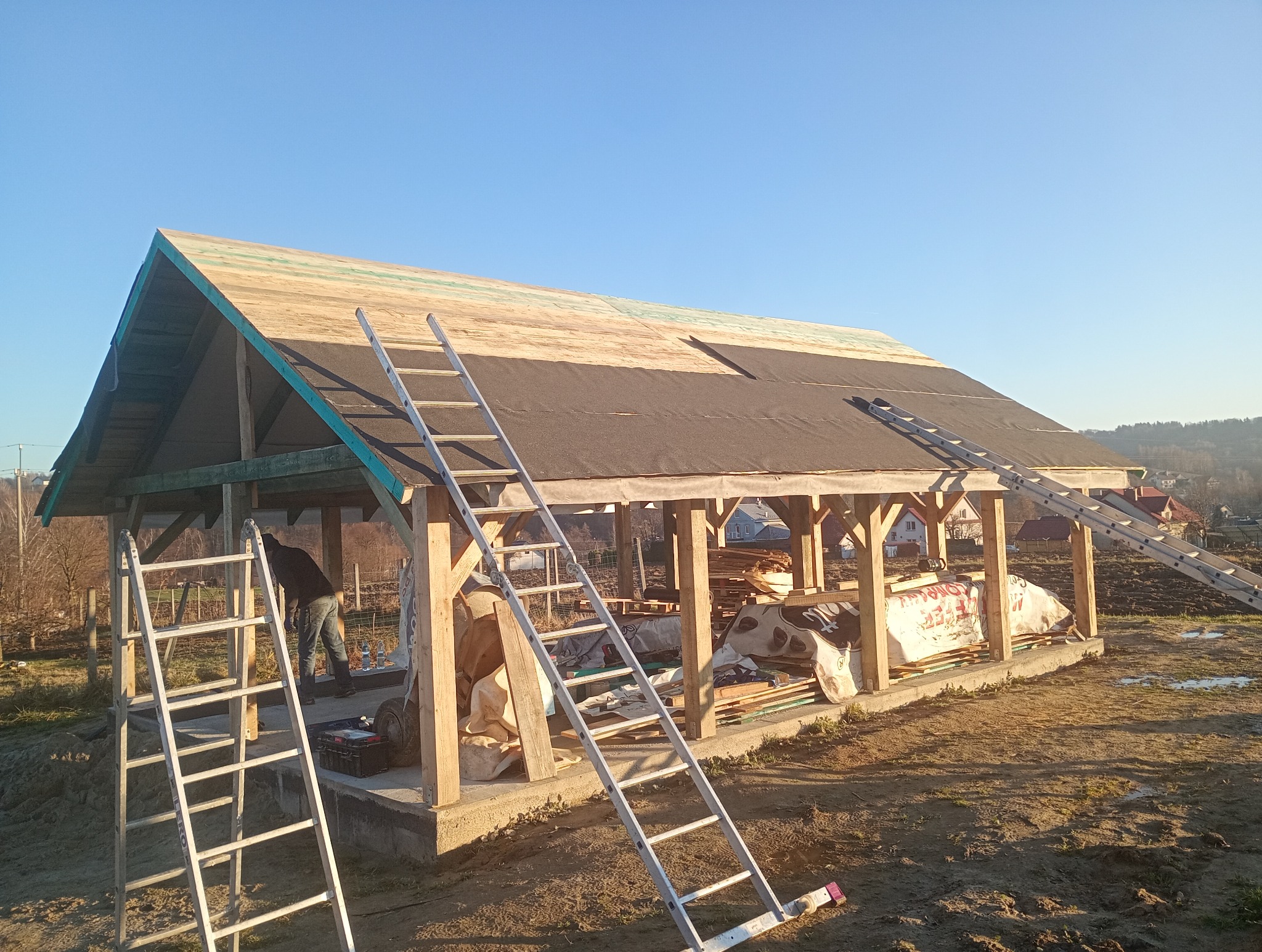Konstrukcja drewniana z częściowo położonym dachem z papy, dwie drabiny opierają się o budowlę, w tle widoczne zabudowania i drzewa, słoneczny dzień.