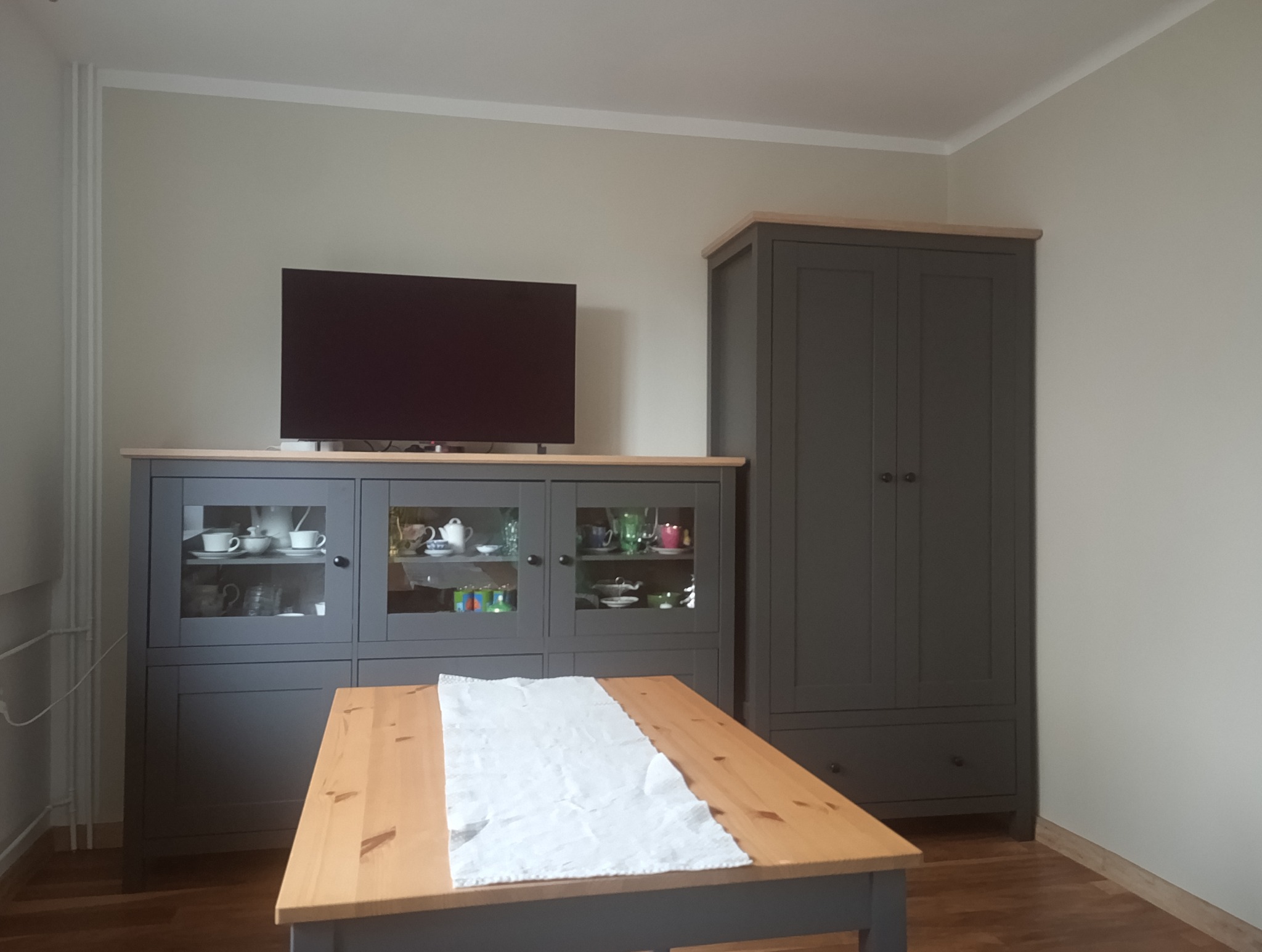Szara szafka z przeszklonymi drzwiczkami, na niej telewizor. Obok wysoka szafa. Na pierwszym planie stół z białym obrusem. Umeblowane wnętrze w stonowanych barwach.