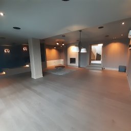 Allremont Kaźmierczak - Przestronne, szare wnętrze po remoncie z drewnianą podłogą, oświetleniem punktowym i kominkiem. Widok na schody w tle. Minimalistyczny design.