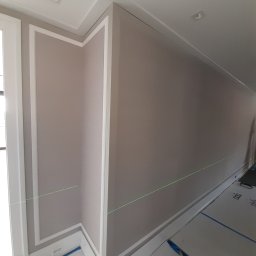 Allremont Kaźmierczak - Ściana w jasnym odcieniu beżu z dekoracyjnymi białymi listwami i laserowym poziomowaniem. Widoczne gniazdka elektryczne i zabezpieczona podłoga.