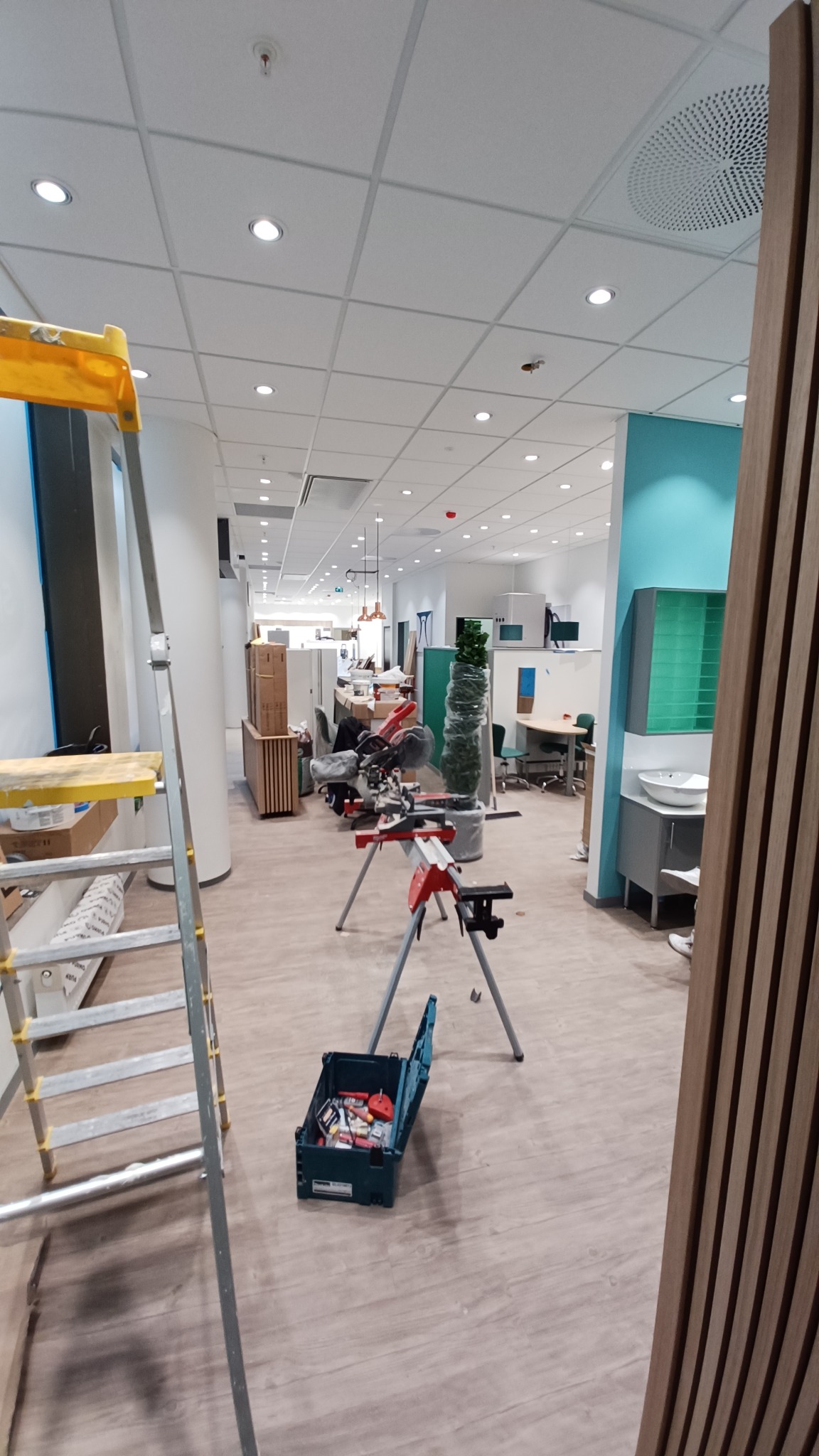 Remont biura: widok na przestrzeń z narzędziami, drabiną i piłą. W tle biurka i elementy wykończenia ścian. Jasne oświetlenie sufitowe.