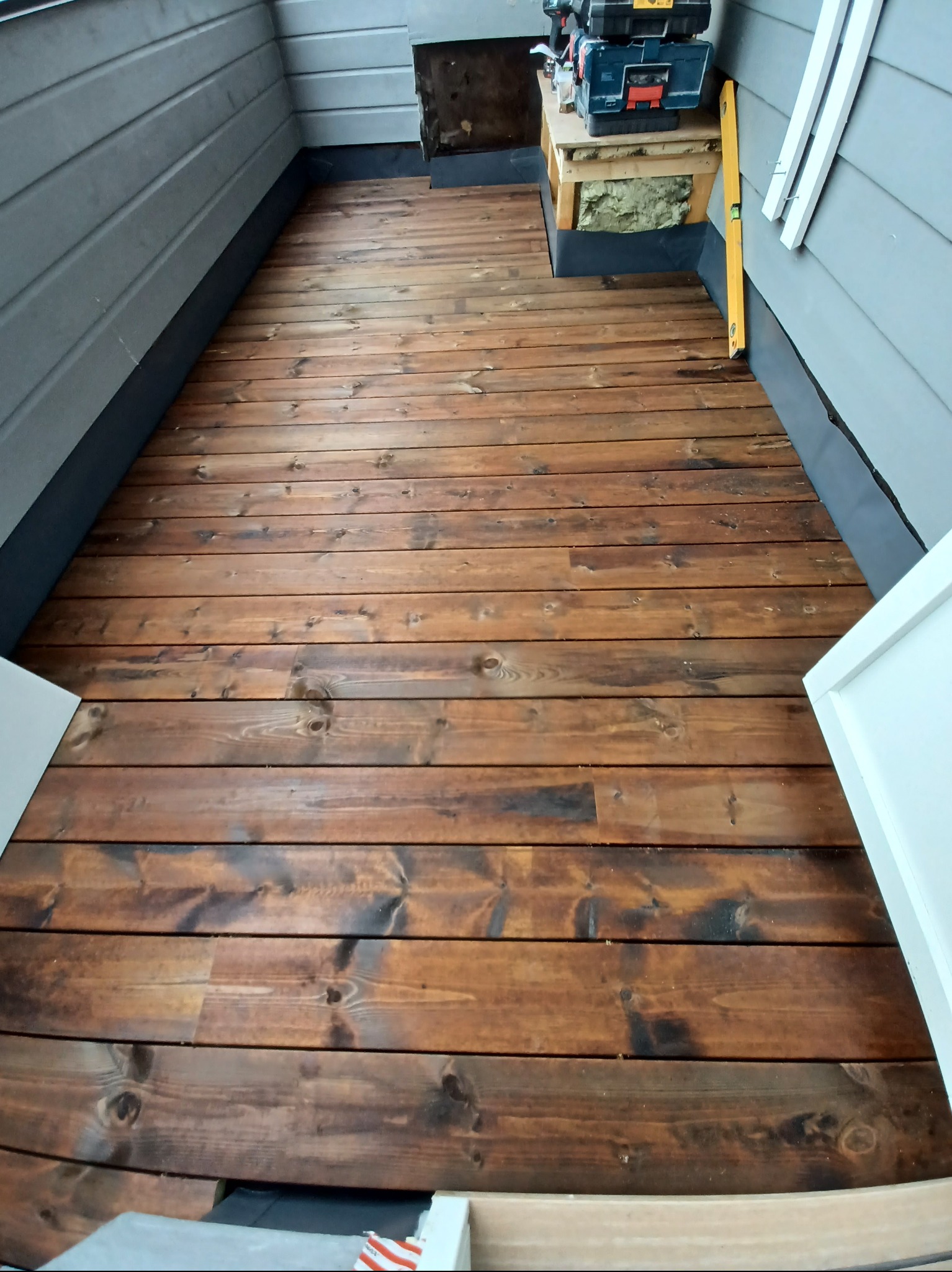 Wykończona podłoga z desek w odcieniach brązu, z widocznymi narzędziami i poziomnicą w tle. Deski ułożone równolegle, tworzą ciepły i naturalny wygląd.