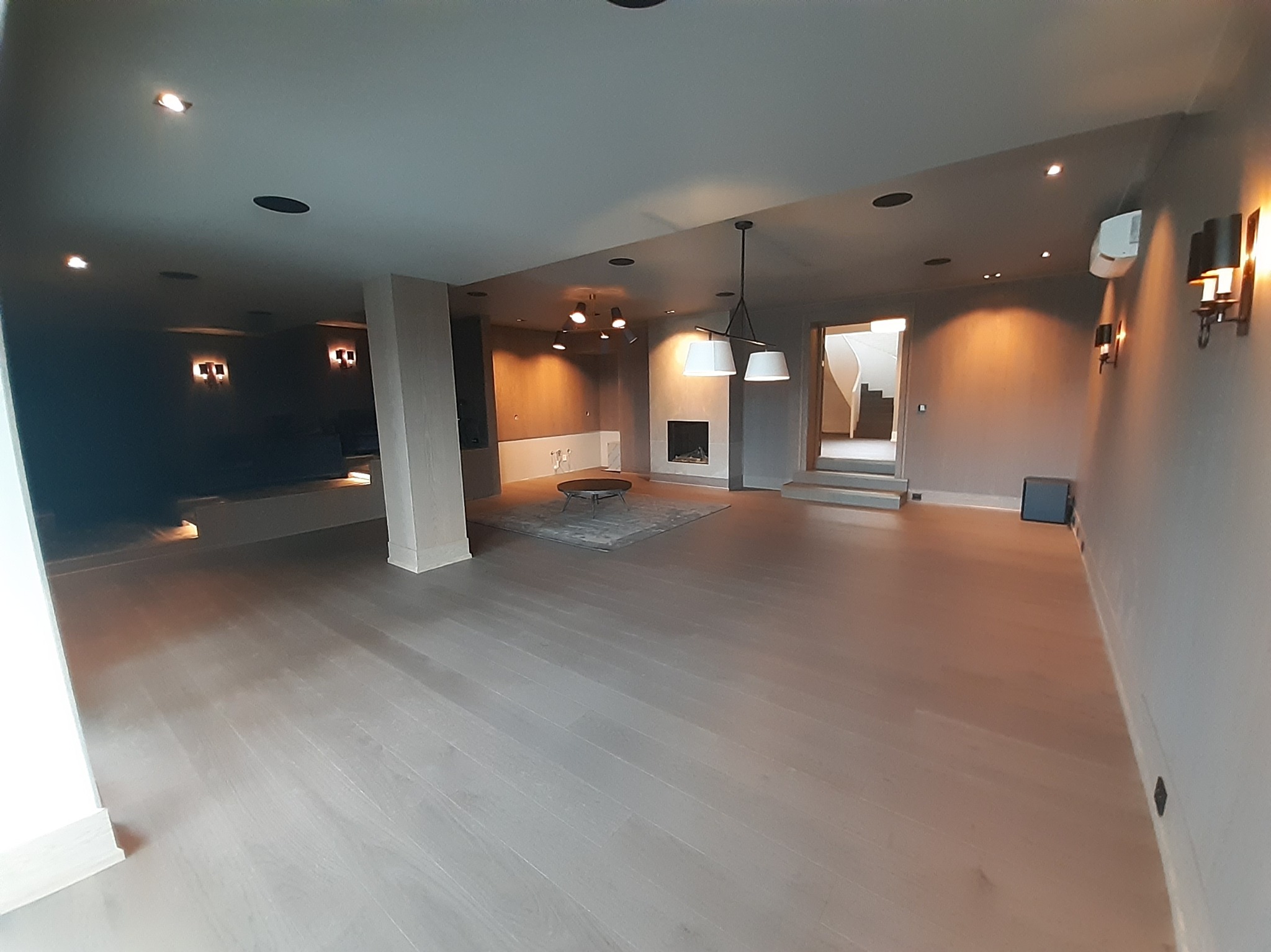 Przestronne, szare wnętrze po remoncie z drewnianą podłogą, oświetleniem punktowym i kominkiem. Widok na schody w tle. Minimalistyczny design.