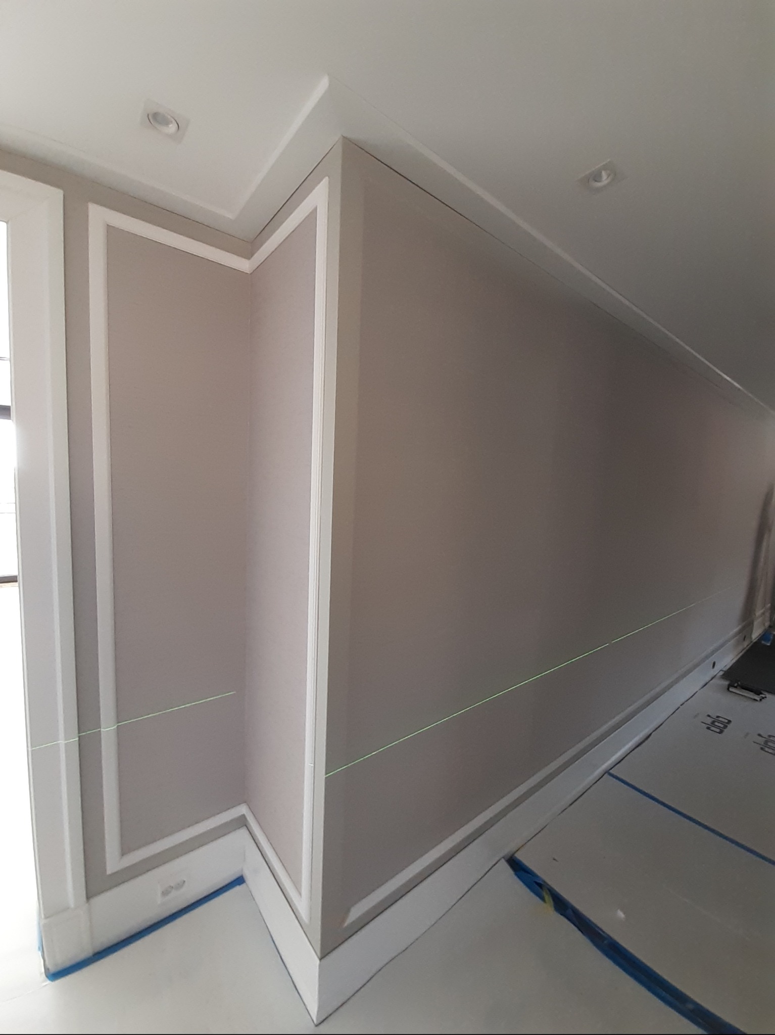 Ściana w jasnym odcieniu beżu z dekoracyjnymi białymi listwami i laserowym poziomowaniem. Widoczne gniazdka elektryczne i zabezpieczona podłoga.