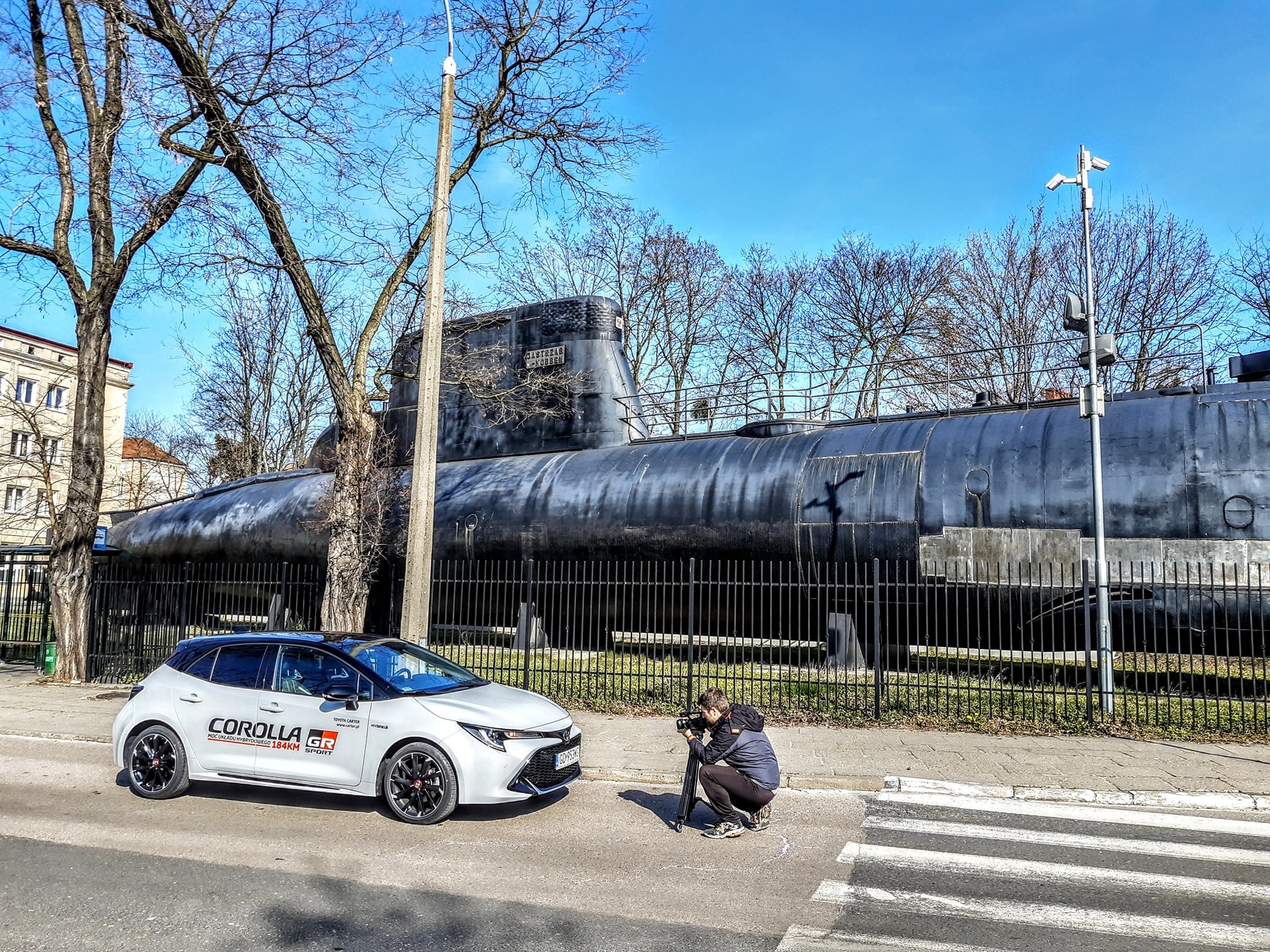 Kamerzysta filmuje białą Toyotę Corollę GR na tle okrętu podwodnego. Ujęcie z niskiej perspektywy, słoneczny dzień, widoczne drzewa bez liści i budynek w tle.