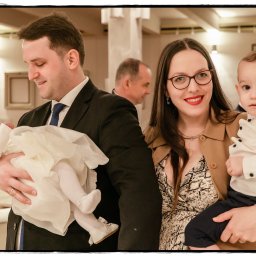 Michał Drewnicki FilmPR - Rodzinne zdjęcie z uroczystości chrztu: rodzice trzymają na rękach dwójkę dzieci, jedno w białej sukience, drugie w koszuli. W tle widoczne elementy wystroju wnętrza.