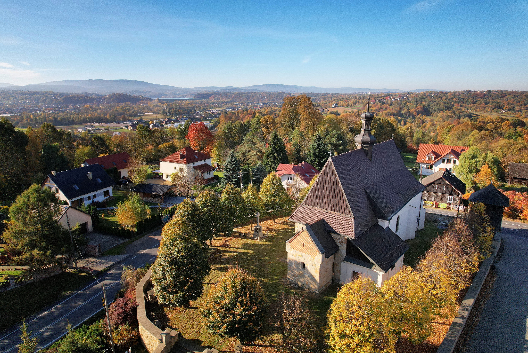 Jesienny krajobraz z lotu ptaka: kościół z ciemnym dachem, domy z czerwonymi dachami, drzewa w odcieniach żółci i czerwieni, góry w oddali pod błękitnym niebem.