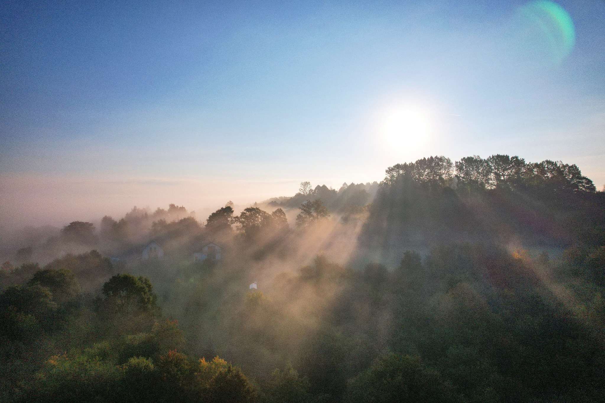 Poranny krajobraz z drona: mgła spowija las i domy, promienie słońca przebijają się przez drzewa, tworząc malowniczy widok. Ujęcie z góry.