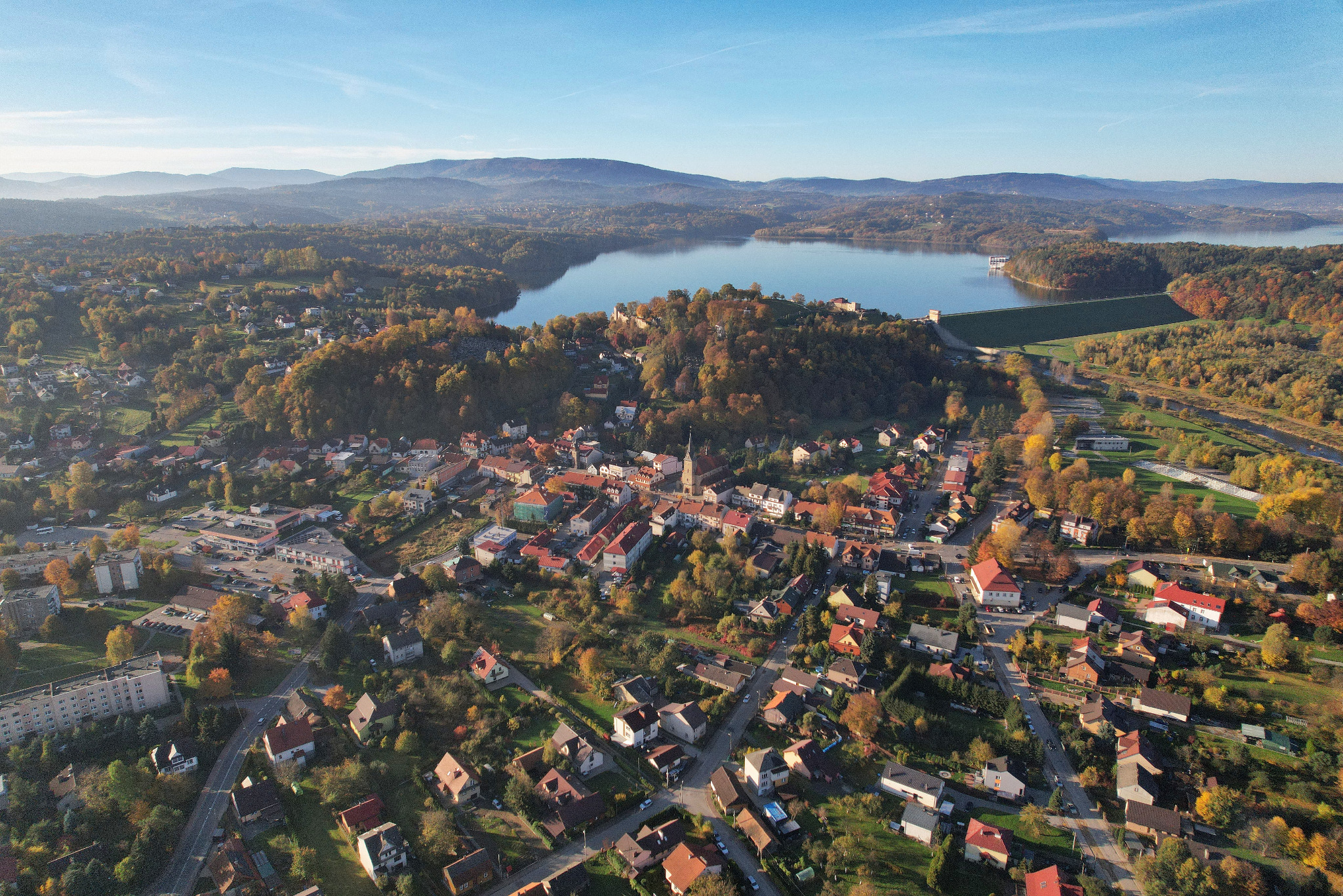 Jesienny krajobraz z lotu ptaka: miasteczko z czerwonymi dachami, otoczone lasami w barwach jesieni, jezioro i góry w tle, wszystko skąpane w porannym słońcu.