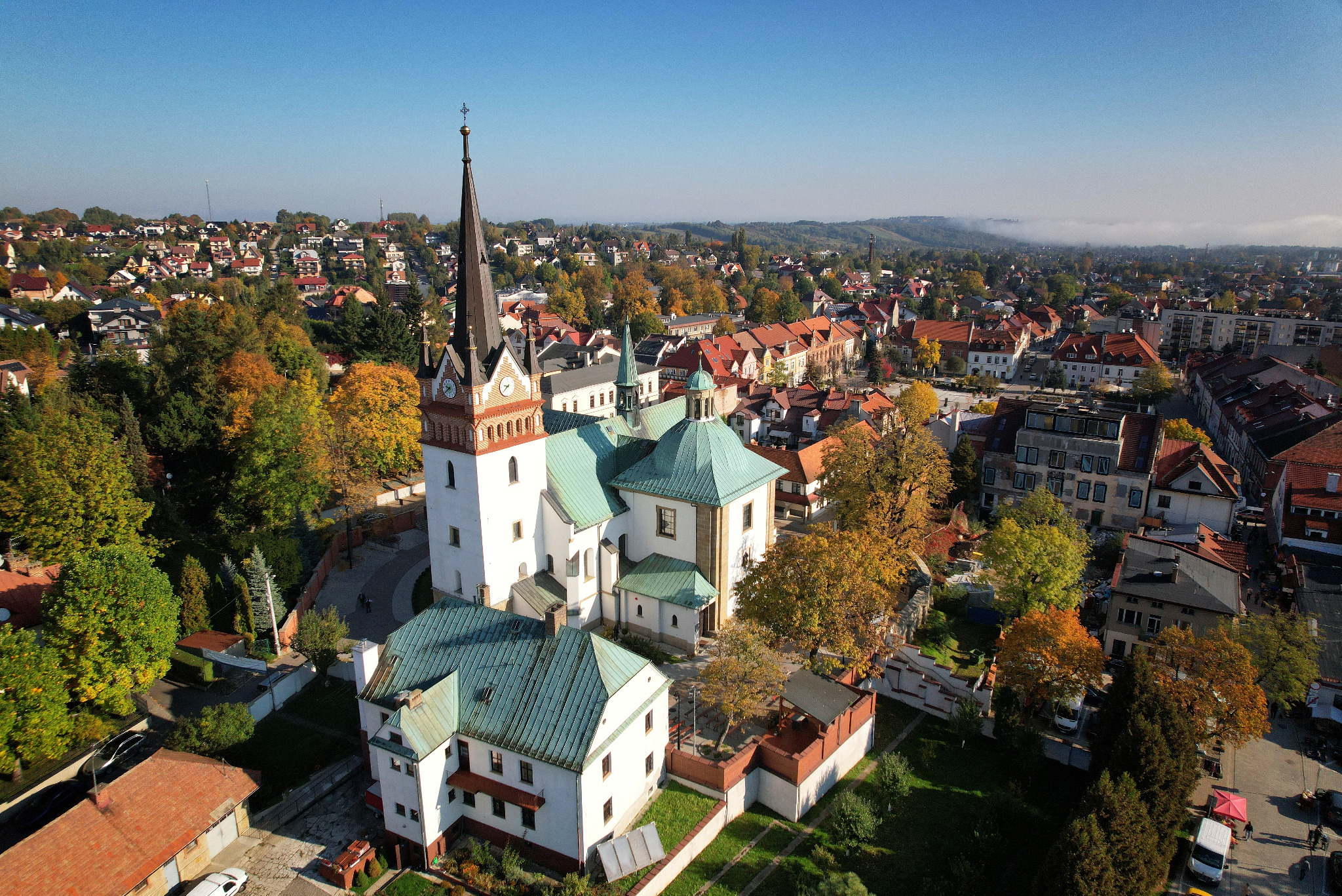Szeroki kadr z drona ukazuje biały kościół z zielonym dachem, otoczony jesiennymi drzewami i zabudową miejską pod błękitnym niebem. Widok na miasto z perspektywy lotu ptaka.
