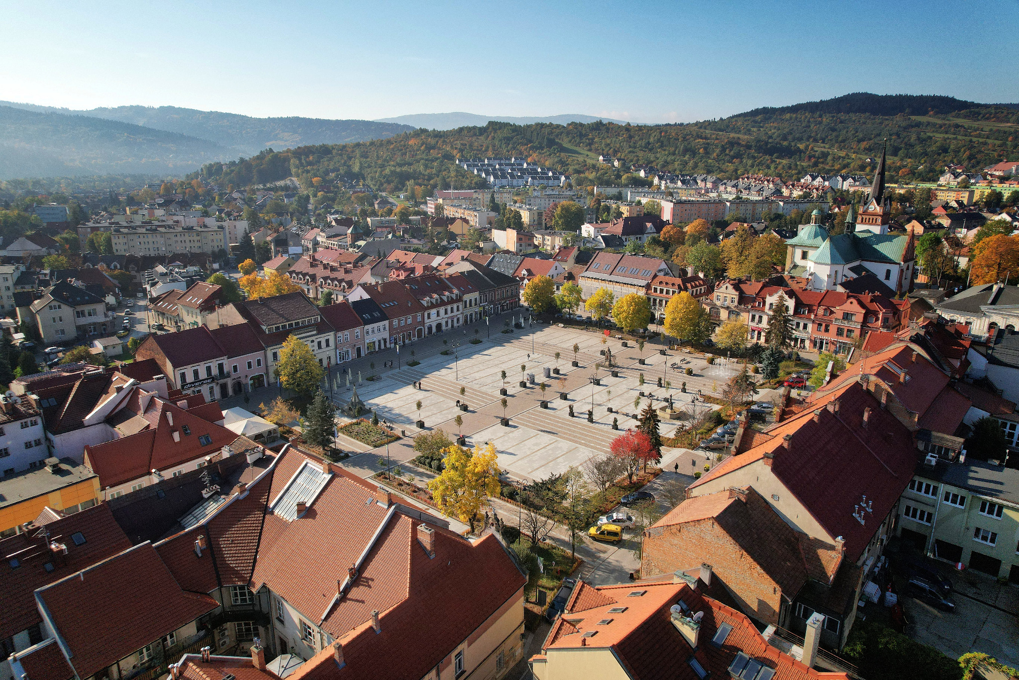 Panoramiczny widok z drona na miasto z rynkiem, czerwone dachy budynków, kościół z zieloną kopułą i góry w tle w jesiennej scenerii.